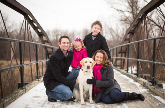 family portrait of five family members and pet dog on a snowy bridge wearing black gray and pink