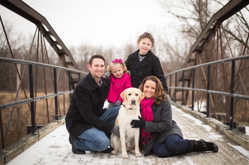 family portrait of five family members and pet dog on a snowy bridge wearing black gray and pink