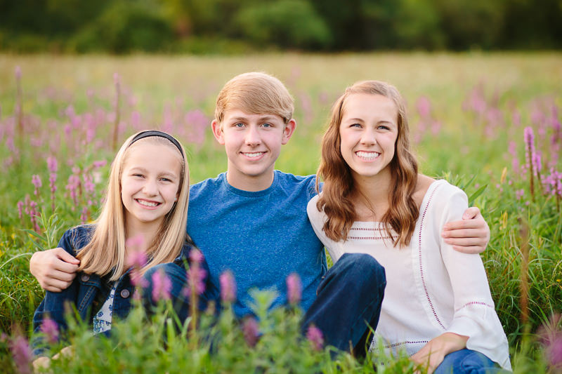 Three happy siblings sitting in grass with purple flowers