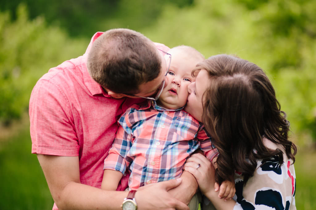 mom and dad kissing baby cheeks in orchard