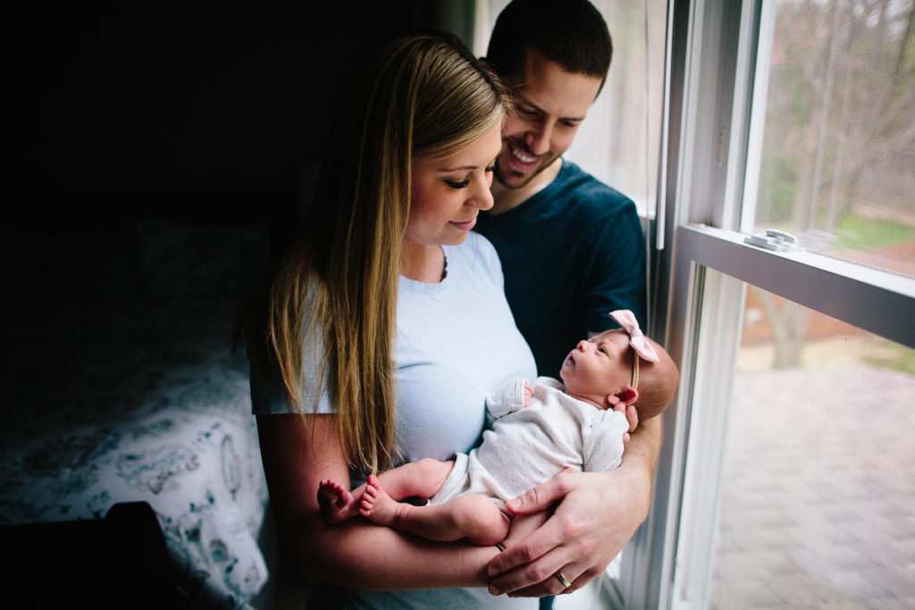mom and dad holding newborn baby girl by window