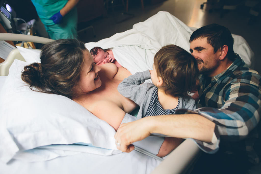 mom smiling at toddler while he checks out his new baby brother