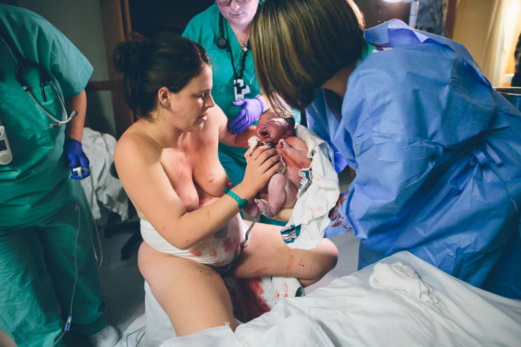 midwife holds baby while mom gets off birth stool umbilical cord still attached