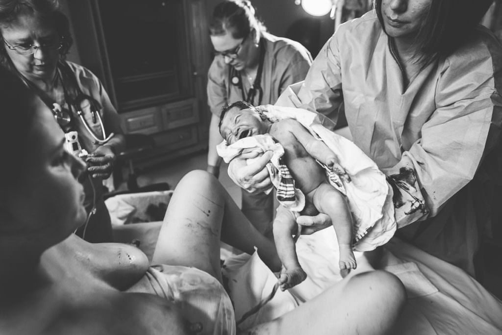 mom lays on bed after giving birth and midwife hand her new baby umbilical chord still attached
