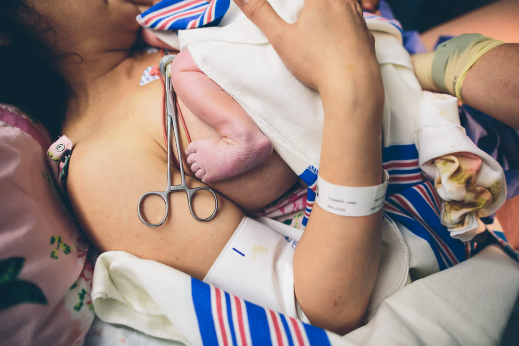 moms arm wrapped around baby on her chest right after birth with baby foot and scissors clamped on umbilical chord