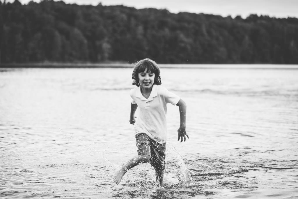 young boy with long brown hair runs through moreau lake fully clothed